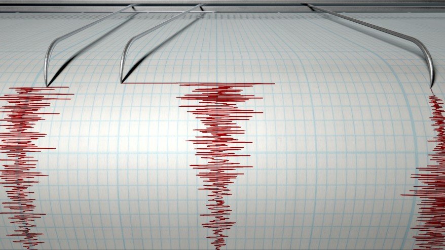 A closeup of a seismograph machine needle drawing a red line on graph paper depicting seismic and eartquake activity 
